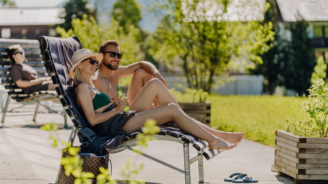 Ein Paar entspannt auf Liegen im Freien, umgeben von Grün. Die Frau trägt einen Sonnenhut und Sonnenbrille, während sie einen Drink genießt. | © Christine Höflehner Photography