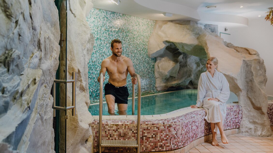 Ein entspannender Wellnessbereich mit einem Innenpool, umgeben von Felsen. Ein Mann steht im Wasser, während eine Frau in einem Bademantel sitzt. | © Christine Höflehner Photography