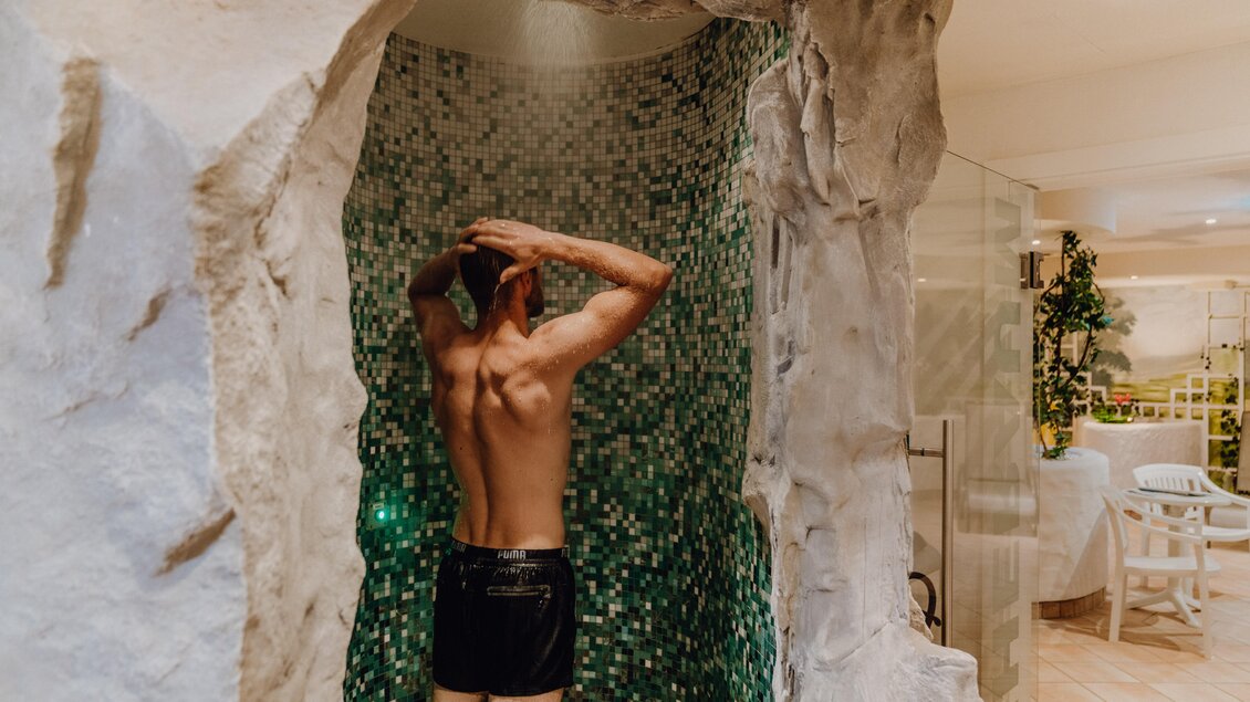 Eine Person steht unter einer Dusche in einem modernen Wellnessbereich. Die Wände sind mit grünen Fliesen und Natursteinen gestaltet. | © Christine Höflehner Photography