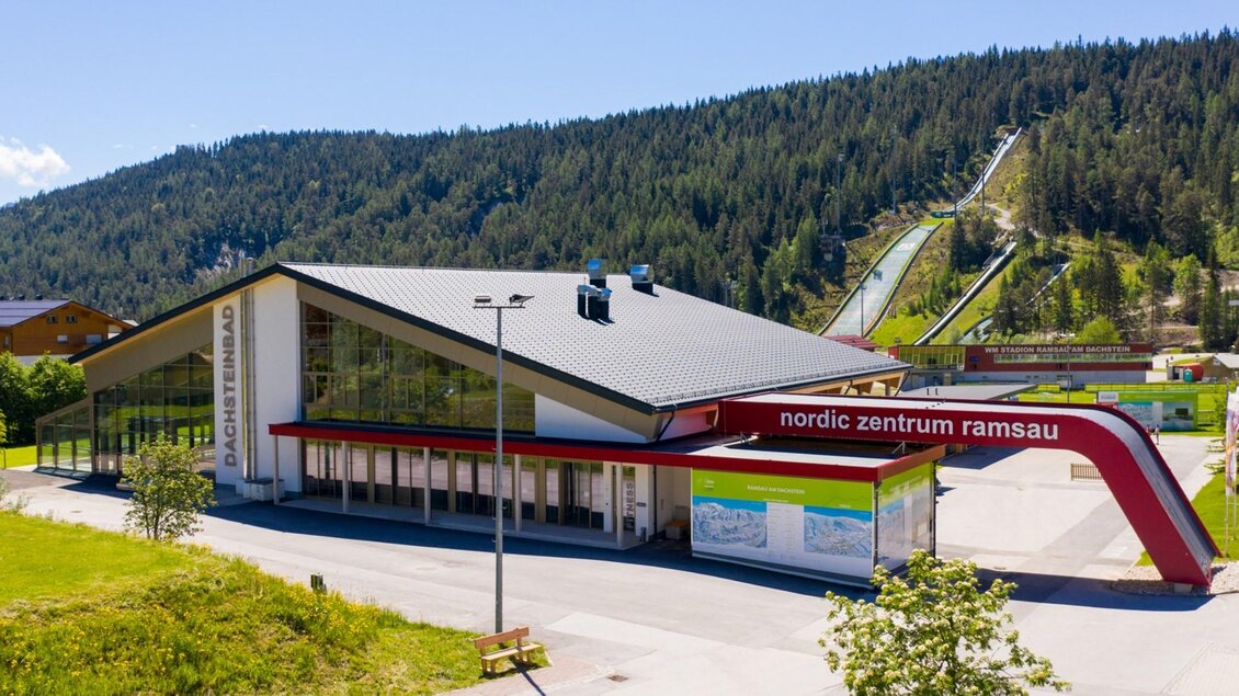 Ein modernes Gebäude des Nordic Zentrums Ramsau umgeben von grünen Wäldern. Im Hintergrund sind Sprungschanzen zu sehen. | © dachsteinbad
