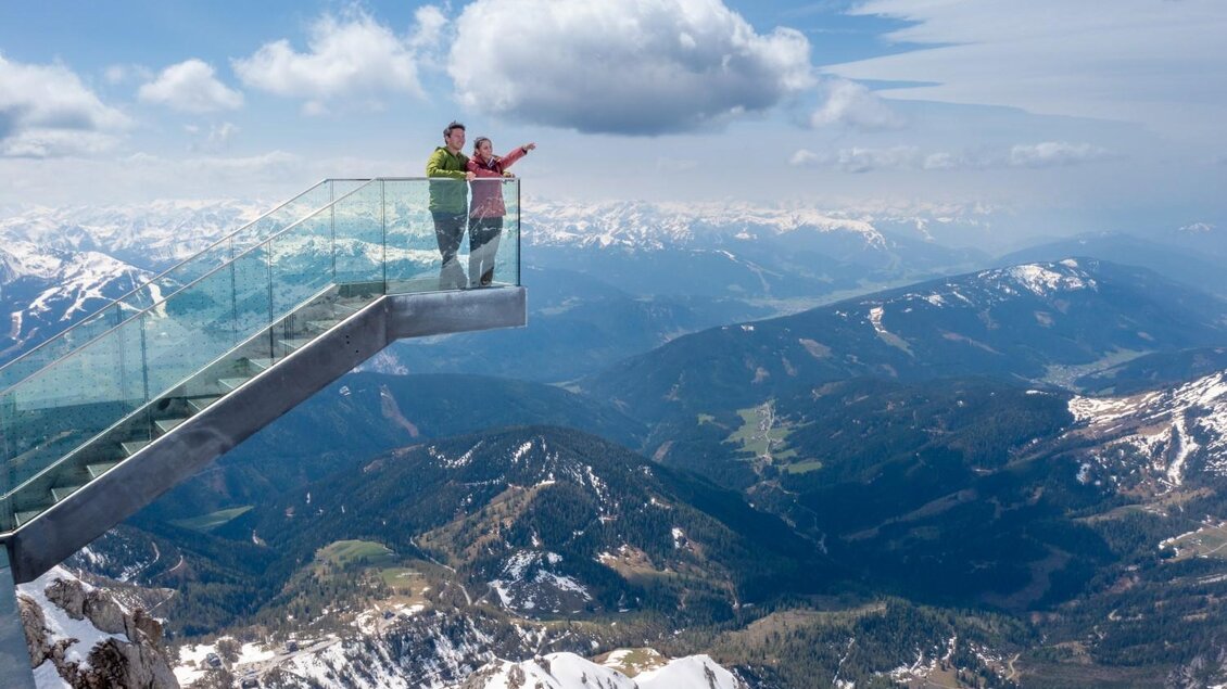Ein Aussichtspunkt auf einem Berg mit Glasgeländer bietet einen atemberaubenden Blick auf die umliegenden Berge und Täler. Zwei Personen zeigen auf die Landschaft und genießen die Aussicht. | © Harald Steiner
