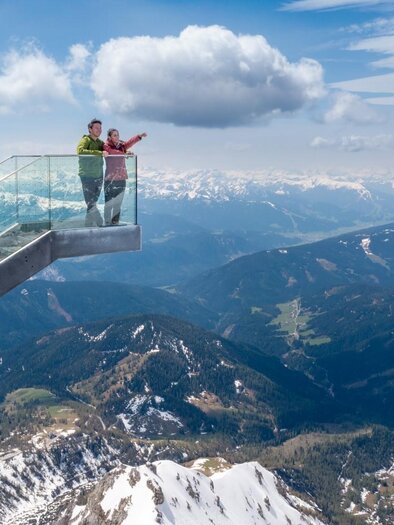 Ein Aussichtspunkt auf einem Berg mit Glasgeländer bietet einen atemberaubenden Blick auf die umliegenden Berge und Täler. Zwei Personen zeigen auf die Landschaft und genießen die Aussicht. | © Harald Steiner