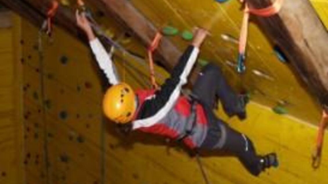 Eine Person klettert an einer Indoor-Kletterwand mit bunten Griffen. Sie trägt einen Helm und Sicherheitsgurte. | © dt-adventure.at
