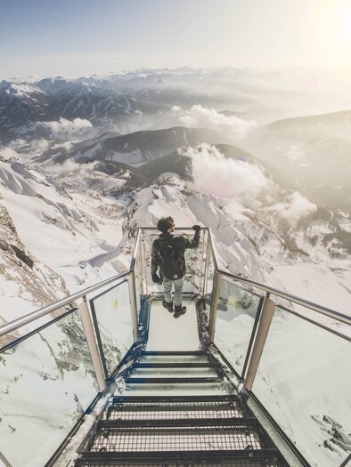 An impressive view from a glass terrace in the high mountains. In the background, snow-capped mountains and a cloudy sky can be seen. | © David McConaghy