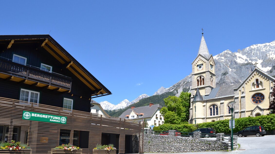 Ein malerisches Dorf mit einem traditionellen Holzhaus und einer beeindruckenden Kirche im Hintergrund. Die Berge sind klar sichtbar, und der Himmel ist strahlend blau. | © tita.at