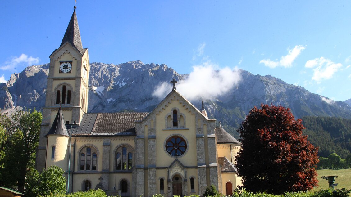 Eine schöne Kirche mit einem hohen Turm und bunten Fenstern, umgeben von grünen Bäumen. Im Hintergrund sind majestätische Berge und ein klarer Himmel sichtbar. | © photo-austria.at/Hans Simonlehner