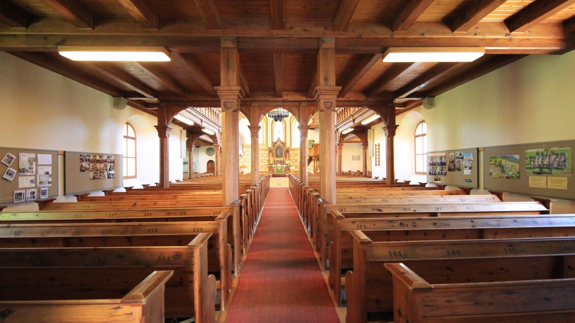 Eine Innenansicht einer Kirche mit Holzbänken und einem langen Teppich. Der Raum zeigt eine ruhige Atmosphäre mit fensterbildlichen Verzierungen.