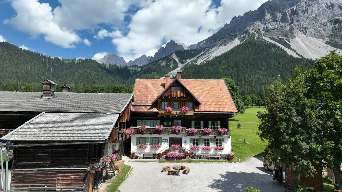 Ein malerisches Holzhaus mit Blumenkästen in einem grünen Tal. Im Hintergrund erheben sich majestätische Berge unter einem blauen Himmel mit Wolken. | © Hans-Peter Steiner