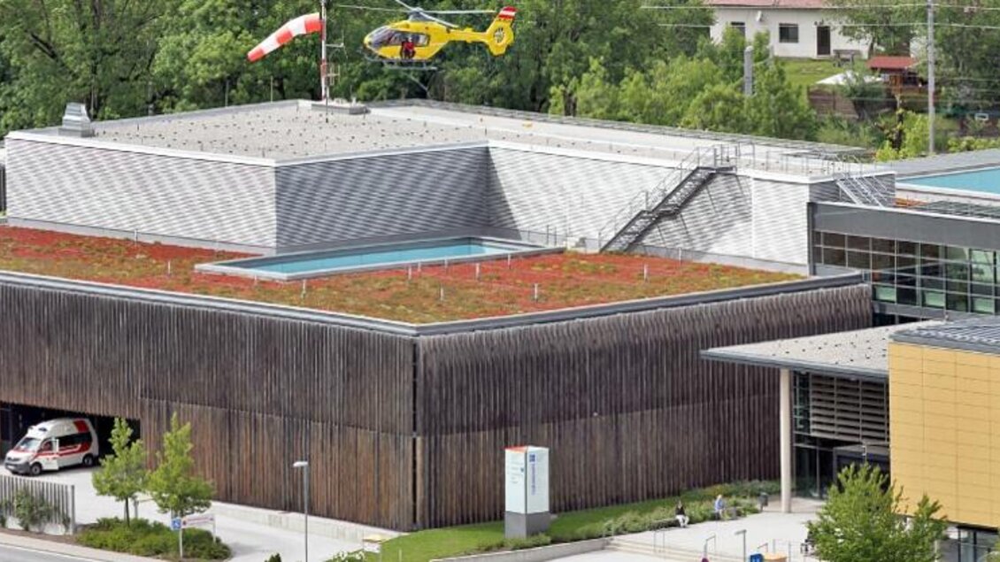 Ein modernes Gebäude mit einem begrünten Dach und einem Hubschrauberlandeplatz. Auf dem Gelände stehen ein Rettungswagen und einige Bäume. | © Christoph Huber