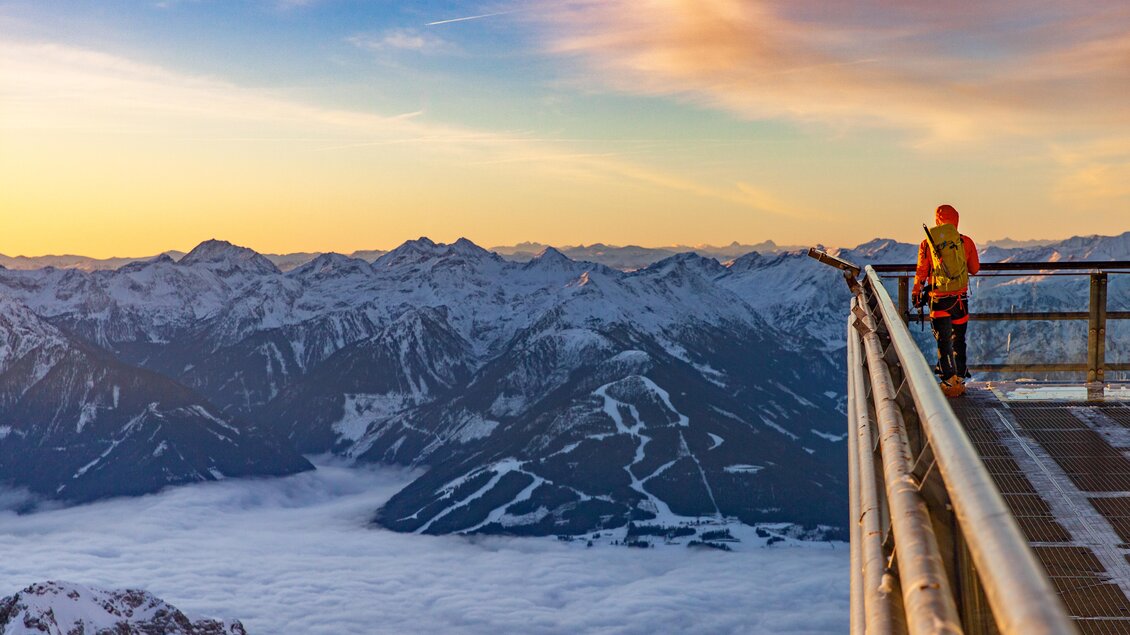 Leicht beschneite Berge ranken aus einem Nebelmeer hervor, während die Sonne im Hintergrund aufgeht. Dabei fotografiert ein Mensch auf einer Aussichtsplattform dieses Naturspektakel. | © Renè Eduard Perhab