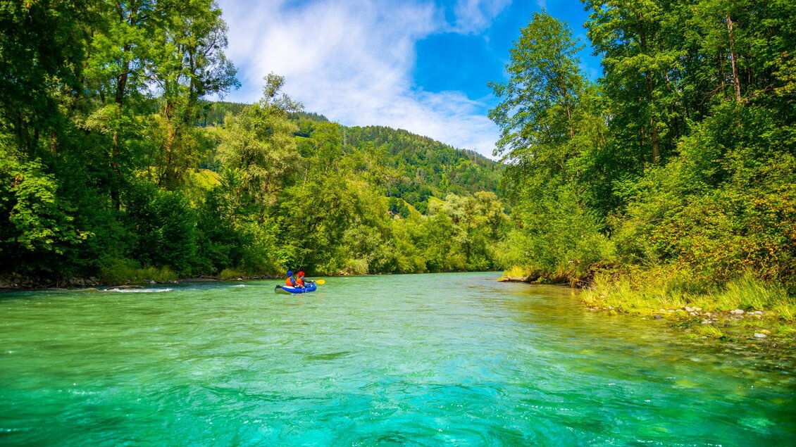Das Bild zeigt eine malerische Flusslandschaft mit üppiger Vegetation an den Ufern und einem blauen Himmel mit vereinzelten Wolken. Ein Kajak mit zwei Personen durchquert das türkisfarbene Wasser des Flusses. | © Kajaktiv