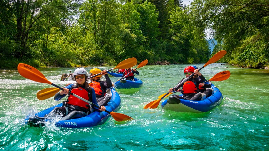 Gruppe von Personen, die an einem sonnigen Tag mit blauen Kajaks paddeln, umgeben von üppigen, grünen Bäumen. Die Kajakfahrer tragen Schutzkleidung und Helme und benutzen orangefarbene Paddel, um sich durch das klare Wasser zu bewegen. | © Kajaktiv