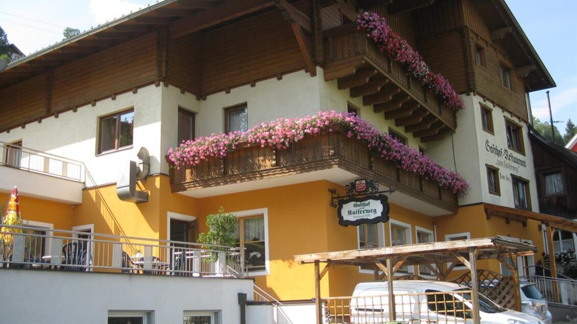 Ein gemütliches Hotel im traditionellen Stil mit blühenden Blumen an den Balkonen. Die Fassade ist hell und einladend, umgeben von einer schönen Landschaft. | © Gasthof zum Kaiserweg