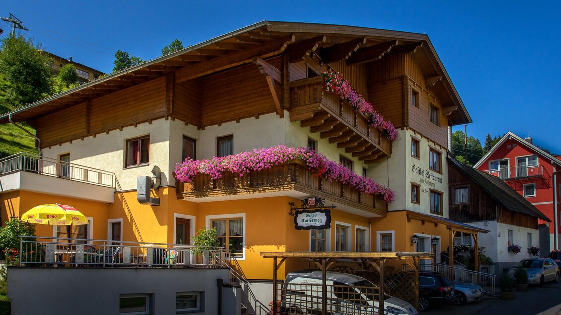 Ein gemütliches Gebäude mit bunten Blumenbalkonen und einem einladenden Außenbereich. Die Architektur ist typisch für alpine Regionen. | © Gasthof zum Kaiserweg