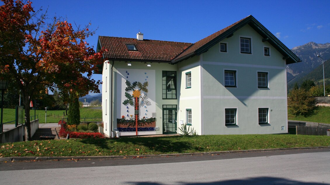 Ein hübsches Gebäude mit einem roten Dach und einer dekorativen Wandmalerei. Umgeben von grünen Wiesen und Bäumen. | © Gemeinde Aich