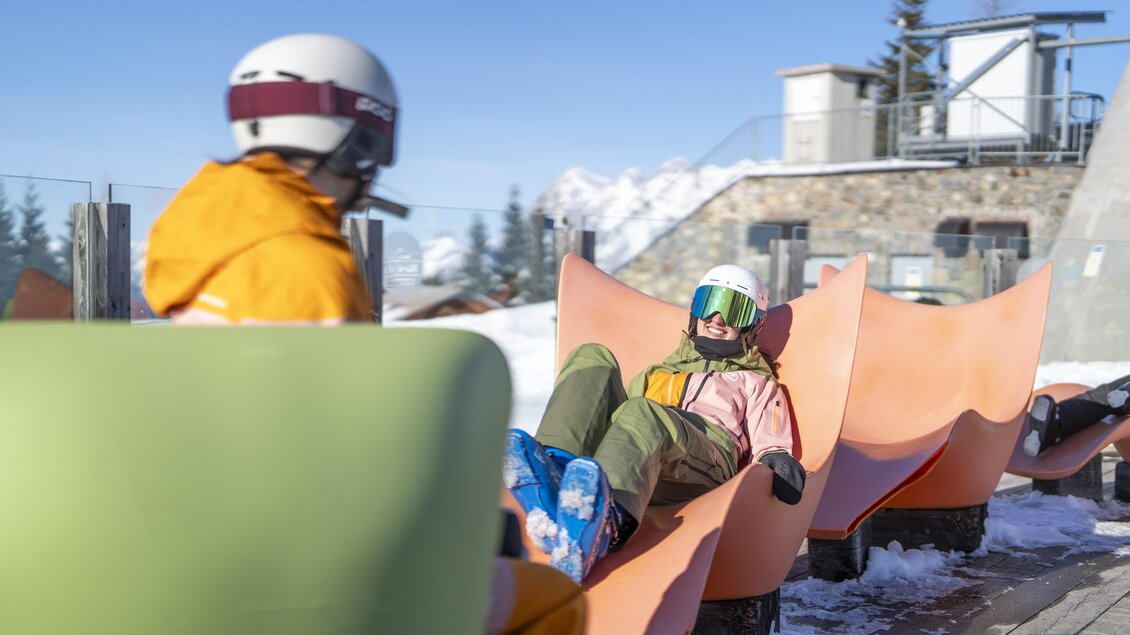 Zwei Personen entspannen auf modernen Sitzgelegenheiten in einer schneebedeckten Berglandschaft. Im Hintergrund sind die verschneiten Gipfel und ein Gebäude sichtbar. | © Hauser Kaibling