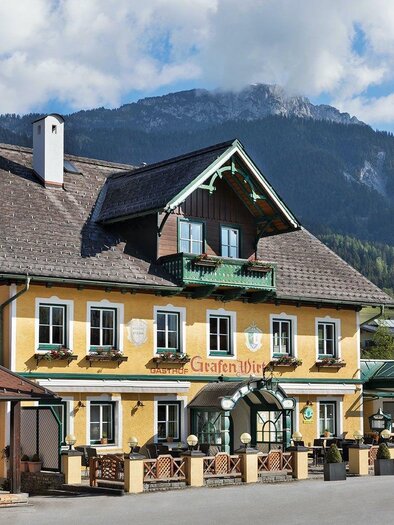 A picturesque inn with a yellow facade and green balcony railing amidst a mountainous landscape. The surroundings are characterized by gentle hills and a clear blue sky. | © GrafenWirt