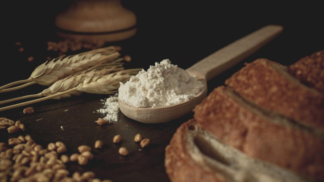 Ein Holzlöffel mit Mehl steht neben frischem Brot und Weizenkörnern. Im Hintergrund sind einige Ähren zu sehen. | © Pixabay
