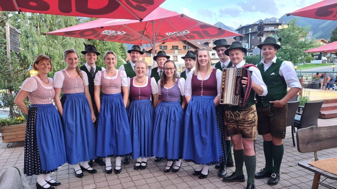 Eine Gruppe von Menschen in traditioneller Tracht steht zusammen, während sie lächeln. Im Hintergrund sind Bäume und Gebäude sichtbar. | © Freistoana zGröbming