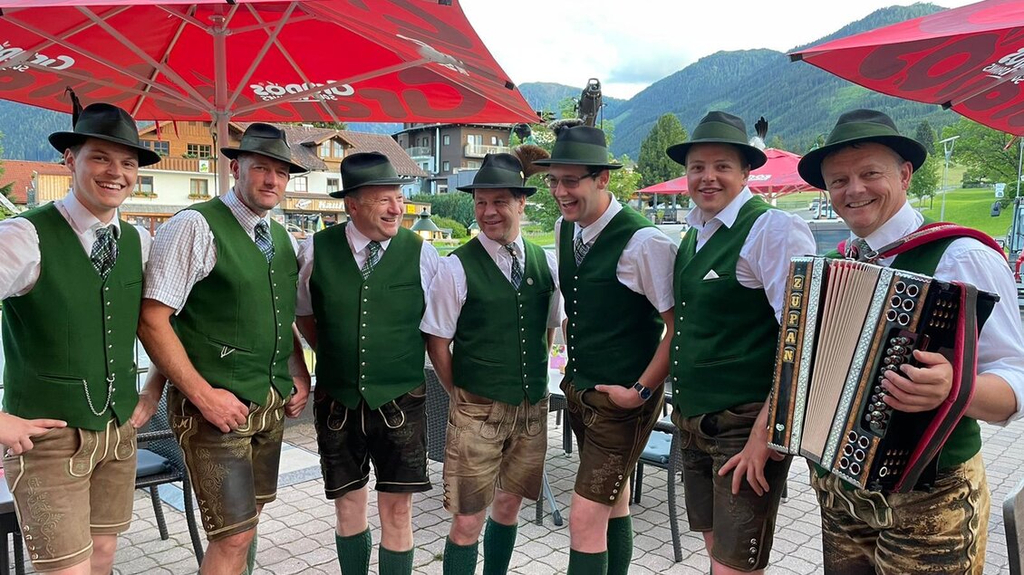 Eine Gruppe von Männern in traditionellen bayerischen Trachten steht zusammen. Sie tragen grüne Westen und Lederhosen und posieren mit einem Akkordeon in der Hand. | © Freistoana zGröbming