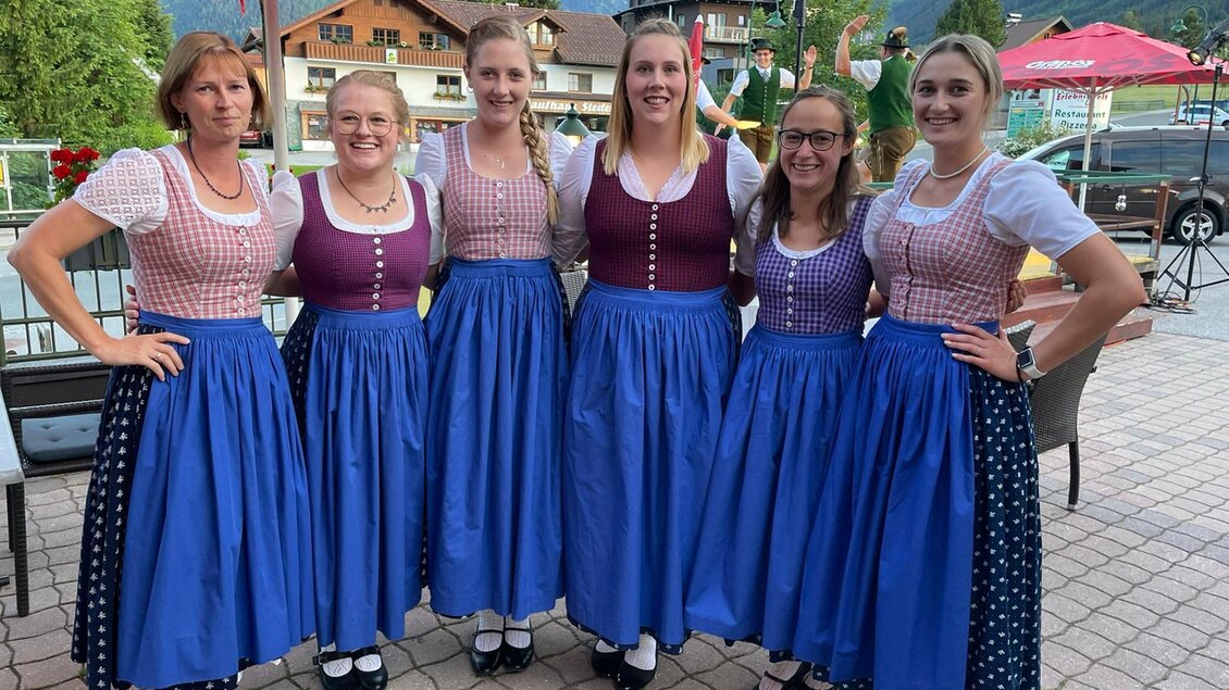 Sechs Frauen in traditionellen Trachten stehen lächelnd zusammen. Im Hintergrund sind Bäume und Pavillons zu sehen. | © FreistoanazGröbming
