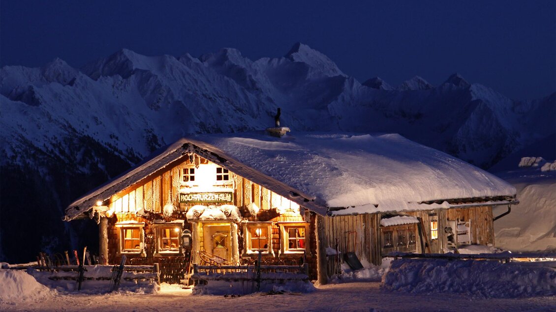 Eine gemütliche Berghütte im Schnee, umgeben von majestätischen Bergen. Die Hütte ist warm erleuchtet und schafft eine einladende Atmosphäre in der Nacht. | © Hochwurzenalm