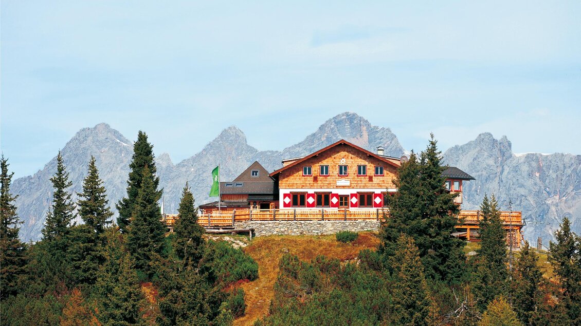 Eine charmante Berghütte umgeben von Tannen und majestätischen Bergen. Der Himmel ist klar und die Aussicht ist beeindruckend. | © Hochwurzenhütte