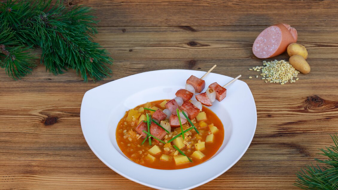 Ein schmackhaftes Gericht in einer weißen Schüssel, bestehend aus einer bunten Suppe mit Kartoffeln und Würstchen. Neben dem Teller liegt eine Wurst und einige Kartoffeln auf einem holzernen Tisch. | © Martin Huber