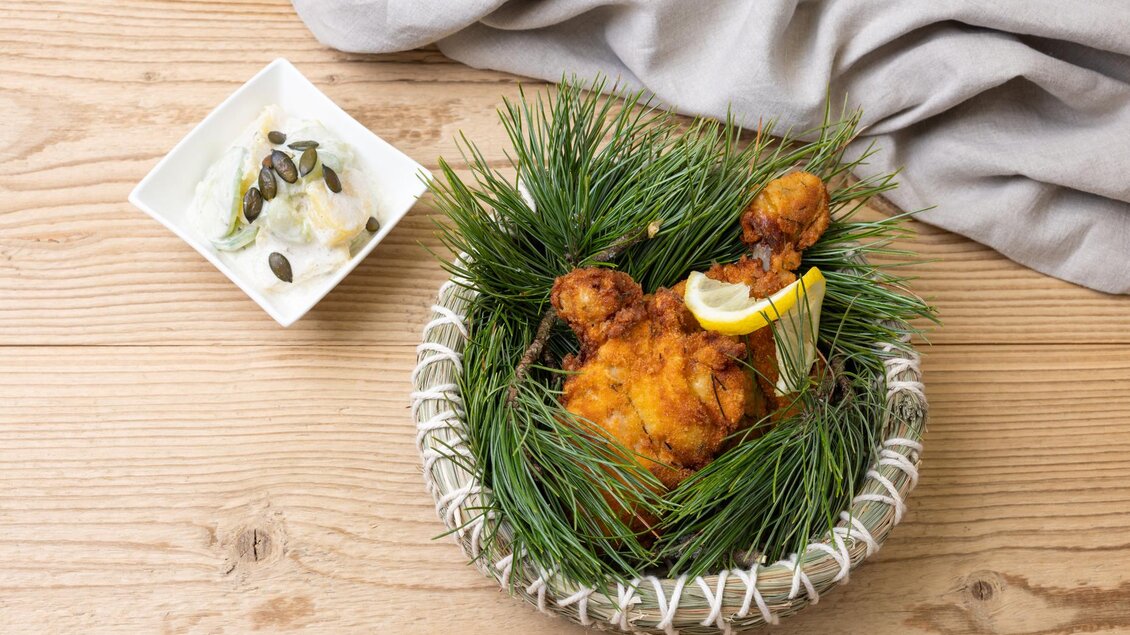 Ein frittiertes Hähnchenstück in einem dekorativen Korb, umgeben von Tannenzweigen. Daneben befindet sich eine kleine Schüssel mit Salat und Kürbiskernen. | © Martin Huber