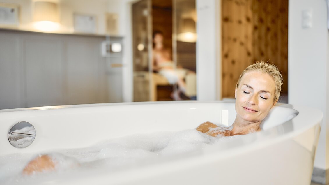 Eine Frau entspannt in einer Badewanne mit Schaum. Im Hintergrund ist eine Sauna zu sehen. | © Thomas Sattler