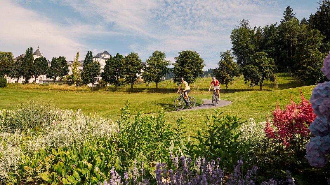 Zwei Radfahrer fahren durch eine grüne Landschaft mit bunten Blumen im Vordergrund. Im Hintergrund sind Bäume und ein Gebäude sichtbar. | © Edy Gröger