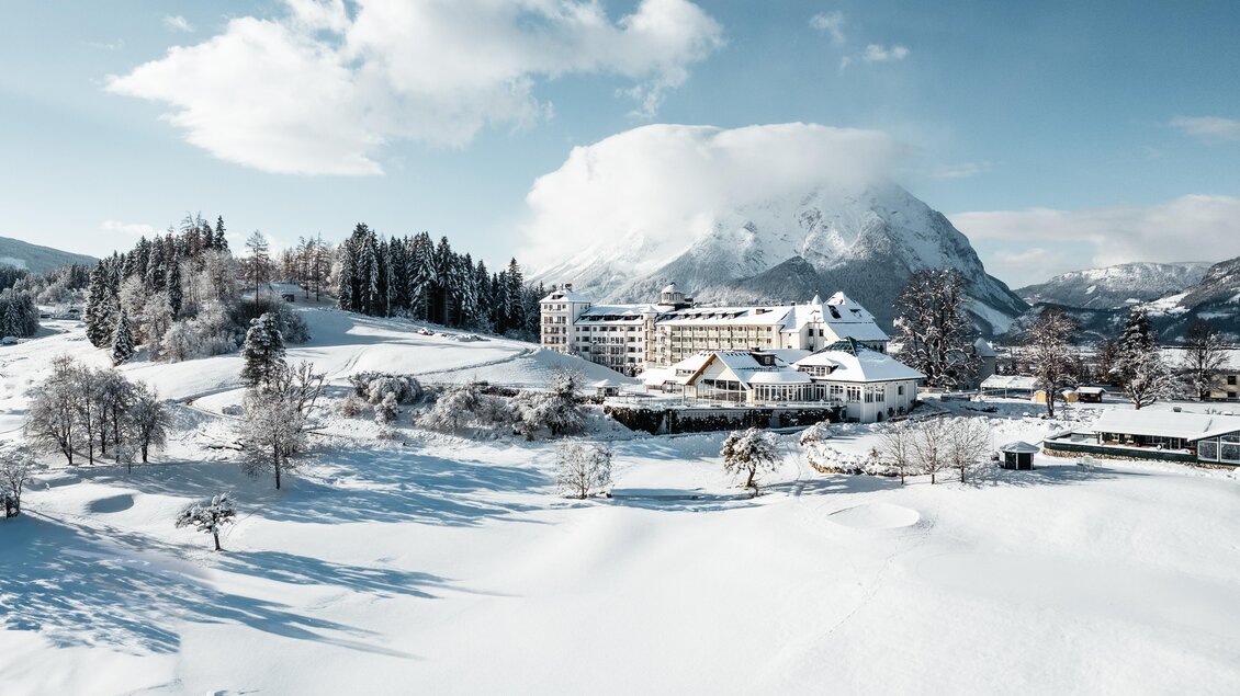 Eine malerische Winterlandschaft mit schneebedeckten Hügeln und einem majestätischen Berg. Im Vordergrund steht ein charmantes Hotel, umgeben von Bäumen und sanften Hügeln. | © Thomas Sattler