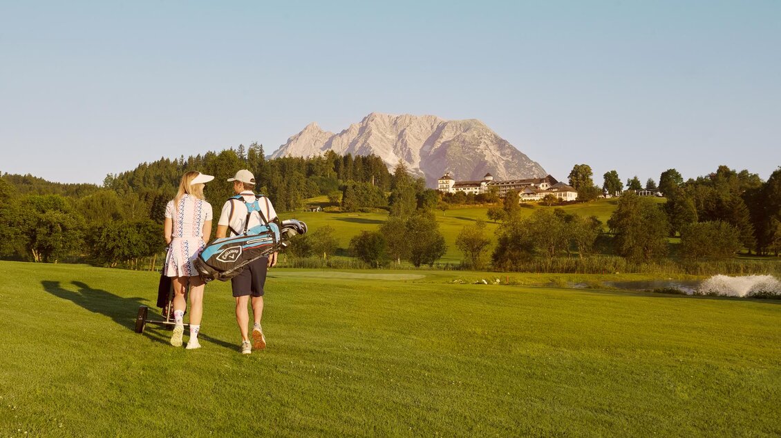 Ein Paar geht über einen Golfplatz mit Blick auf die Berge. Im Hintergrund sind Bäume und ein schönes Anwesen zu sehen. | © Art in action