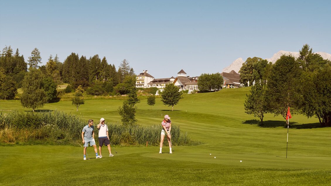 Eine Gruppe von drei Personen spielt Golf auf einer grünen Wiese. Im Hintergrund sind Bäume und ein großes Gebäude zu sehen. | © Art in action