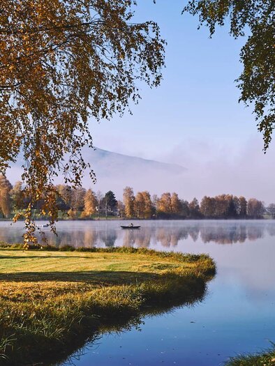 A tranquil lake surrounded by colorful autumn trees. The sky is clear, and the mist lies gently over the water. | © Infobüro Irdning
