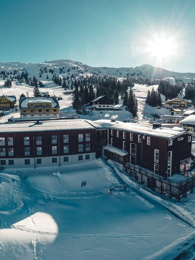 Eine verschneite Berglandschaft mit einem Hotel und einigen kleinen Chalets. Die Sonne scheint über die Berge und die umliegende Natur.