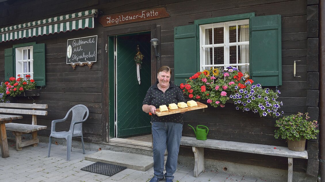 Ein lächelnder Mann steht vor einem traditionellen Holzhaus und hält ein Tablett mit frischen Brötchen. Vor dem Haus sind blühende Blumen und eine einladende Sitzgelegenheit. | © Herfried Marek