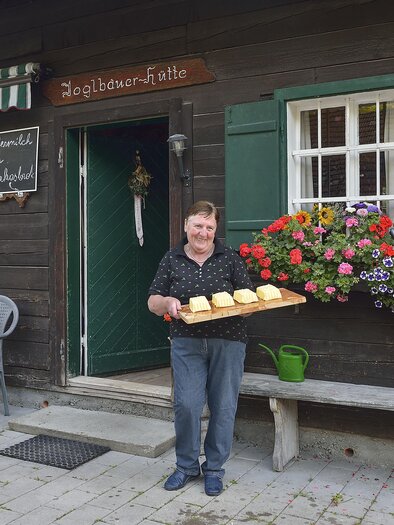Ein lächelnder Mann steht vor einem traditionellen Holzhaus und hält ein Tablett mit frischen Brötchen. Vor dem Haus sind blühende Blumen und eine einladende Sitzgelegenheit. | © Herfried Marek