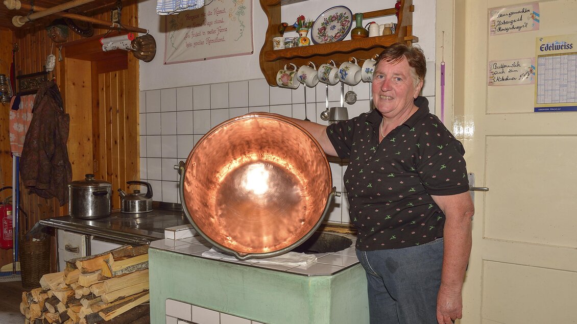 Eine Person steht in einer gemütlichen Küche und hält einen großen Kupfertopf. Im Hintergrund sind Holzscheite und Küchengeräte zu sehen. | © Herfried Marek