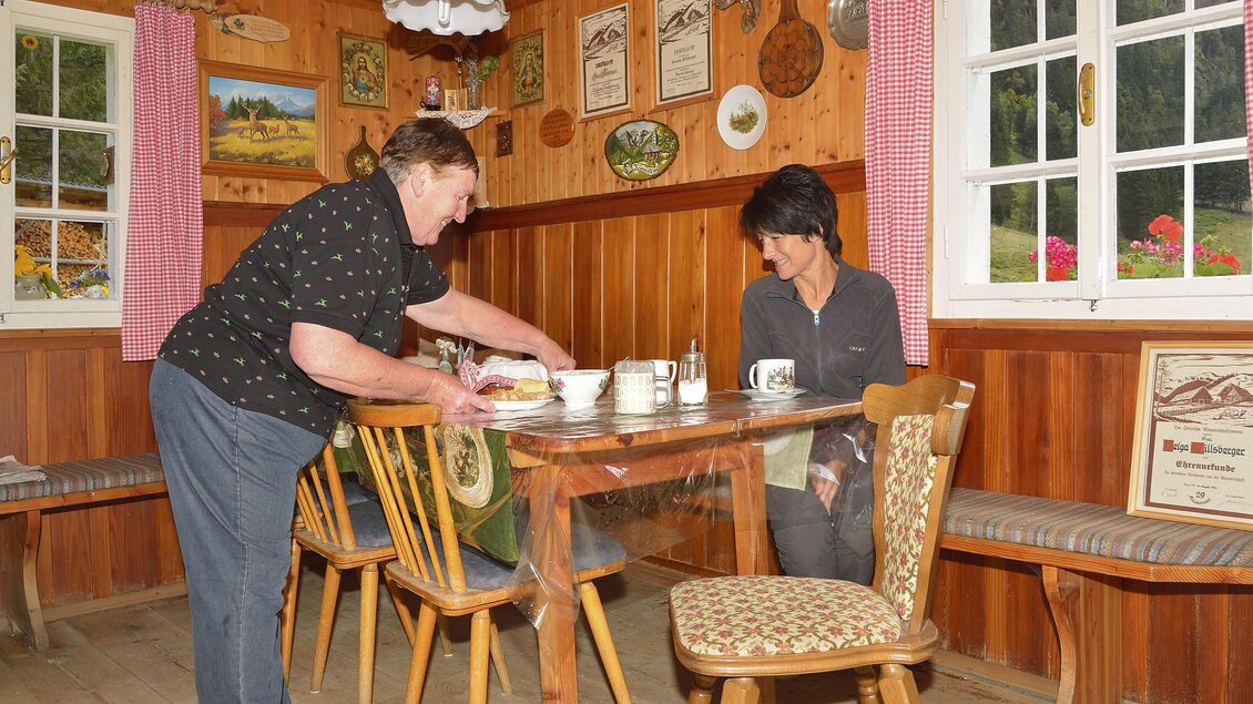 Eine gemütliche Stube mit Holzvertäfelung und traditionellen Dekorationen. Zwei Frauen sitzen am Tisch, während eine von ihnen Geschirr vorbereitet. | © Herfried Marek