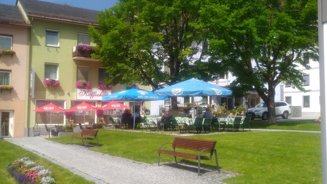 Ein ruhiger Außenbereich mit grüner Rasenfläche und einem Weg. Im Hintergrund stehen Bäume und ein Restaurant mit bunten Schirmen. | © Peer Dieter
