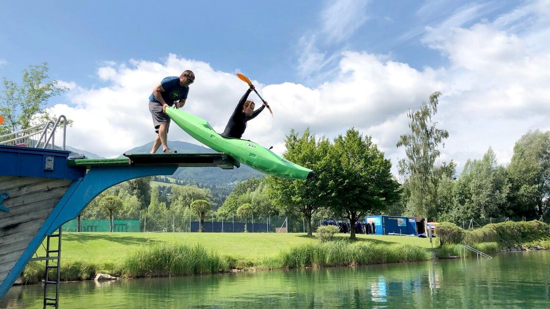 Zwei Personen springen von einem Steg ins Wasser, eine in einem grünen Kajak. Die Umgebung ist grün und sonnig mit einem klaren Himmel.
