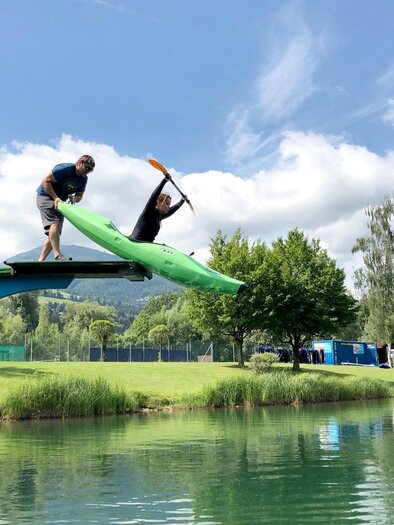 Two people are jumping from a dock into the water, one in a green kayak. The surroundings are green and sunny with a clear sky.