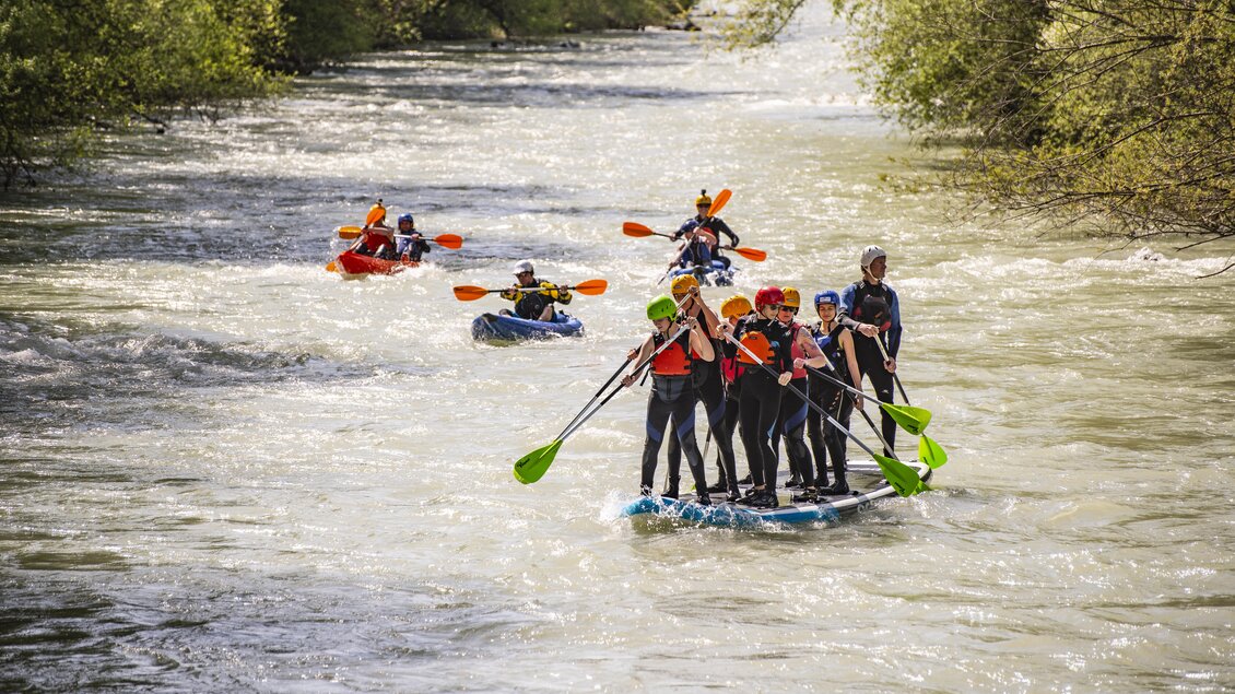 Eine Gruppe von Menschen paddelt auf einem Stand-Up-Paddle-Board auf einem Fluss. Im Hintergrund sind weitere Kajakfahrer zu sehen.