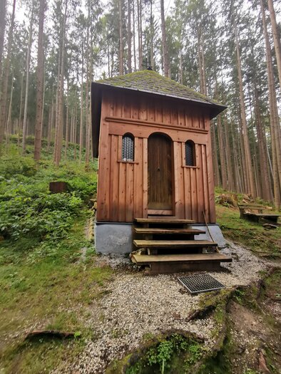 A small, rustic cottage in the woods, surrounded by tall trees and dense vegetation. The front door is made of wood and there are a few steps leading up to the door. | © TVB Schladming-Dachstein