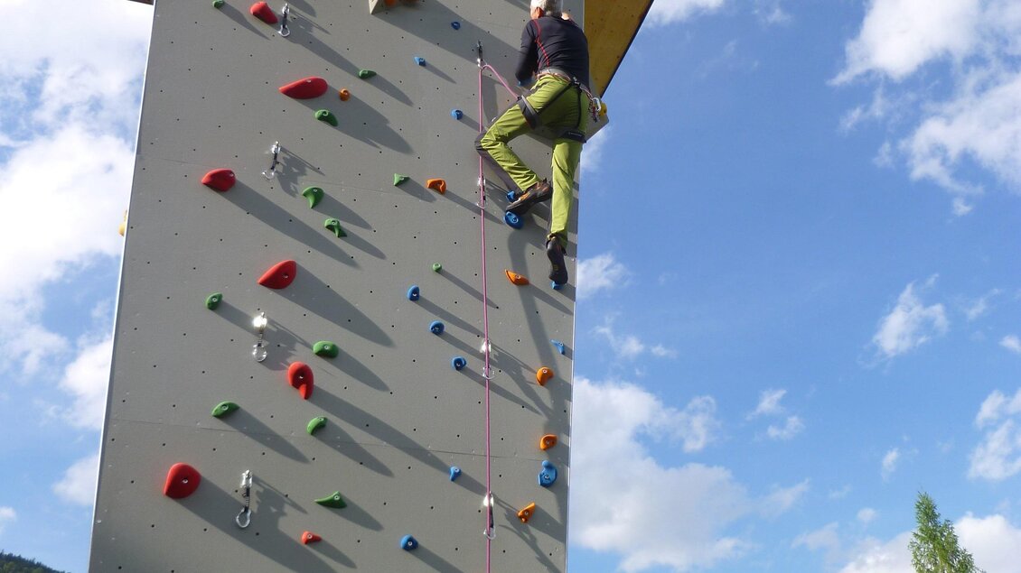 Ein Kletterer erklimmt eine hohe Kletterwand im Freien. Im Hintergrund sind grüne Bäume und ein blauer Himmel mit Wolken zu sehen. | © Gemeinde Aich