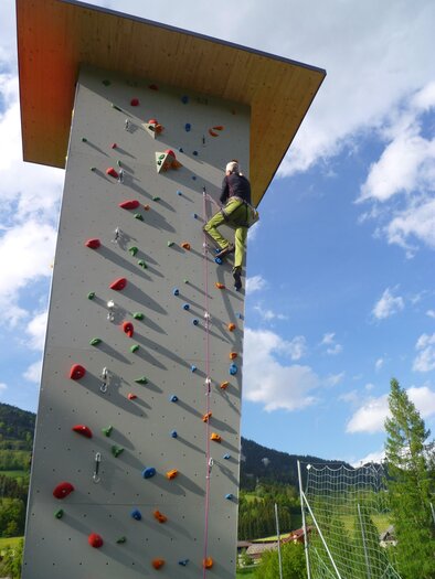Ein Kletterer erklimmt eine hohe Kletterwand im Freien. Im Hintergrund sind grüne Bäume und ein blauer Himmel mit Wolken zu sehen. | © Gemeinde Aich