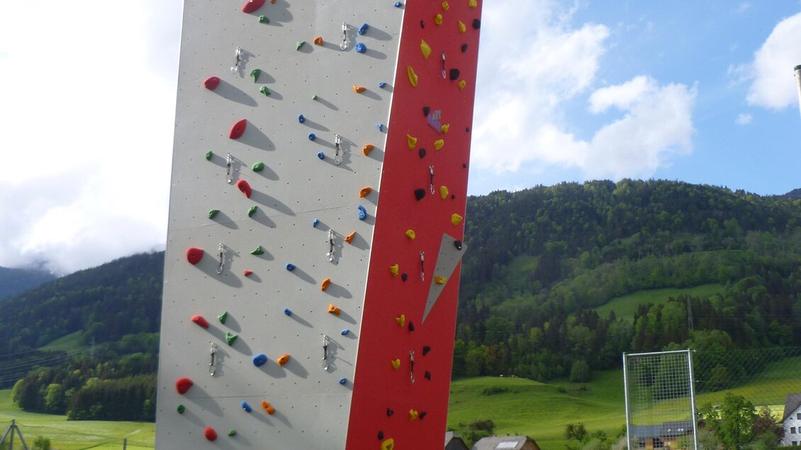 Eine Kletterwand steht in einer grünen Landschaft unter einem blauen Himmel. Im Vordergrund ist ein Kletterer zu sehen, der sich auf das Klettern vorbereitet. | © Gemeinde Aich