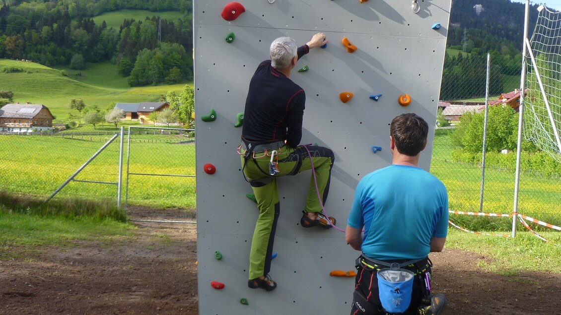 Ein Kletterer steigt an einer Kletterwand, während ein Sicherer daneben steht. Die Umgebung ist grün und ländlich, mit Bergen im Hintergrund. | © Gemeinde Aich