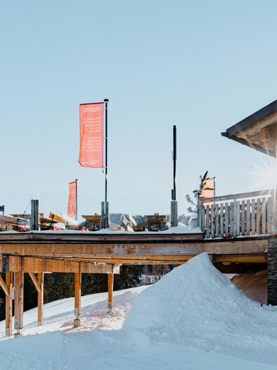 A modern restaurant on a mountain with a wooden terrace and white snowdrifts. The sun shines through the windows while orange flags flutter. | © Knapplhütte